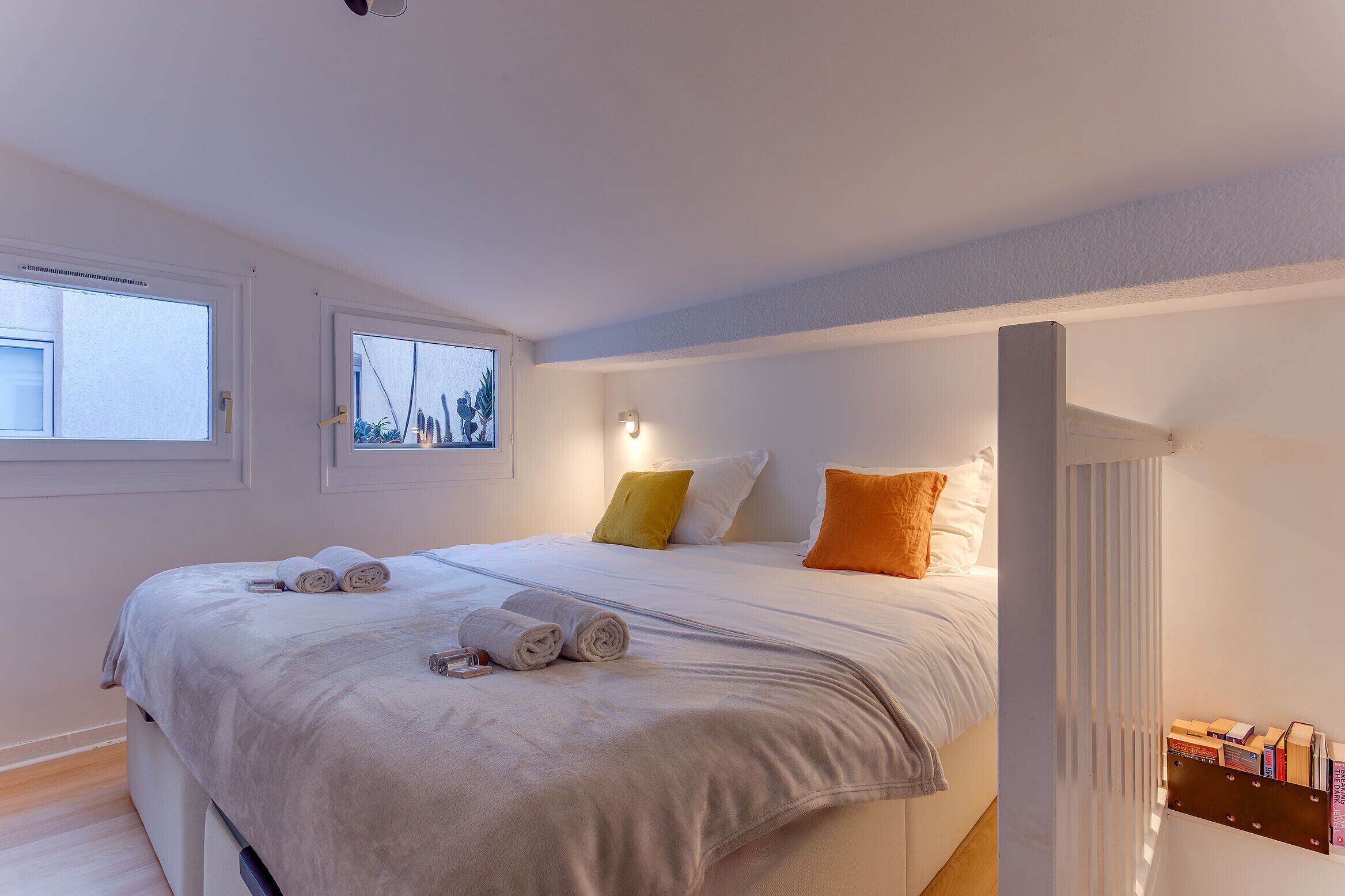 A neatly made bed with rolled towels, a bedside table with books, and a window with a view of trees.