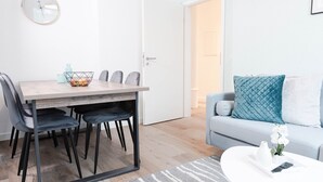 Un soggiorno moderno con zona pranzo, che include un tavolo in legno, sedie rivestite in grigio, un divano bianco con cuscini blu e bianchi e un piccolo vaso bianco con fiori su un tavolino da caffè bianco.