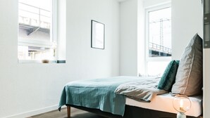 Una camera da letto con un letto, due finestre, un comodino con una lampada e un quadro appeso al muro.