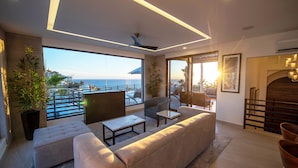 Un salon moderne doté d’une grande fenêtre donnant sur un balcon avec vue sur la mer. On y trouve des canapés, une table basse et un lampadaire.