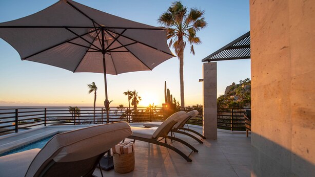 Un espace aménagé au bord de la piscine, avec des chaises longues, un grand parasol et une vue sur le coucher de soleil sur l’océan.