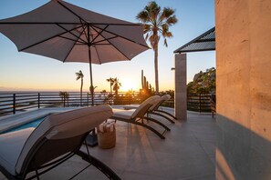 A poolside area with lounge chairs, a large umbrella, and a view of the sunset over the ocean.