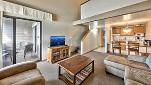 A spacious living area with a sofa, a coffee table, and a TV on a wooden cabinet. There is a sliding glass door leading to an outdoor seating area. A kitchen is visible in the background with wooden cabinets and a chandelier.
