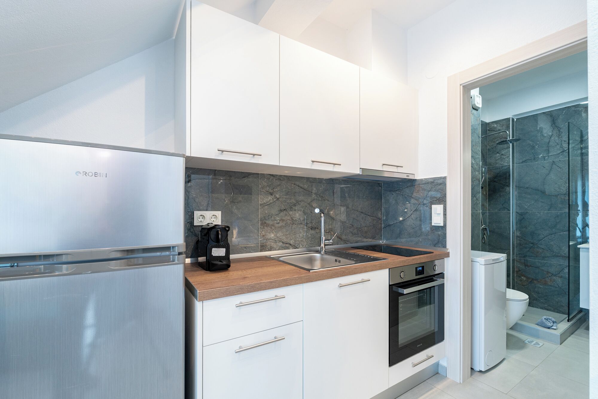 Una cocina moderna con armarios blancos, un refrigerador de acero inoxidable, una cafetera y un horno empotrado.