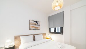 A modern hotel room with a large bed, bedside tables, a wall-mounted lamp, a painting, and a window with a blind. - Erato Residential Complex (Ofrinion)