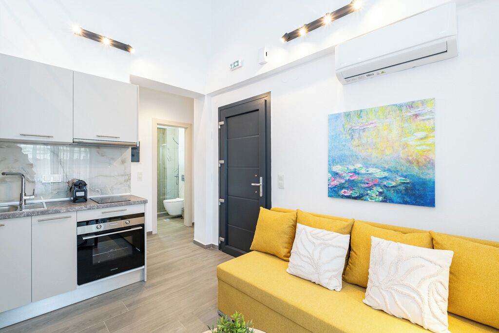 Eine moderne Küche mit weißen Schränken, einem Einbauofen und einer Arbeitsplatte. Ein gelbes Sofa mit weißen Kissen und ein Blumenbild an der Wand.