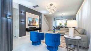 A modern living room with a flat-screen TV, blue velvet armchairs, a beige sofa, a round coffee table, and a dining area in the background. - Lake Shore 5BR Resort Villa • Pool • Game Loft (Davenport)