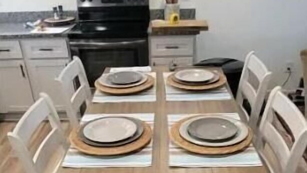 Un comedor con una mesa de madera dispuesta para cuatro personas, sillas blancas y una cocina al fondo.