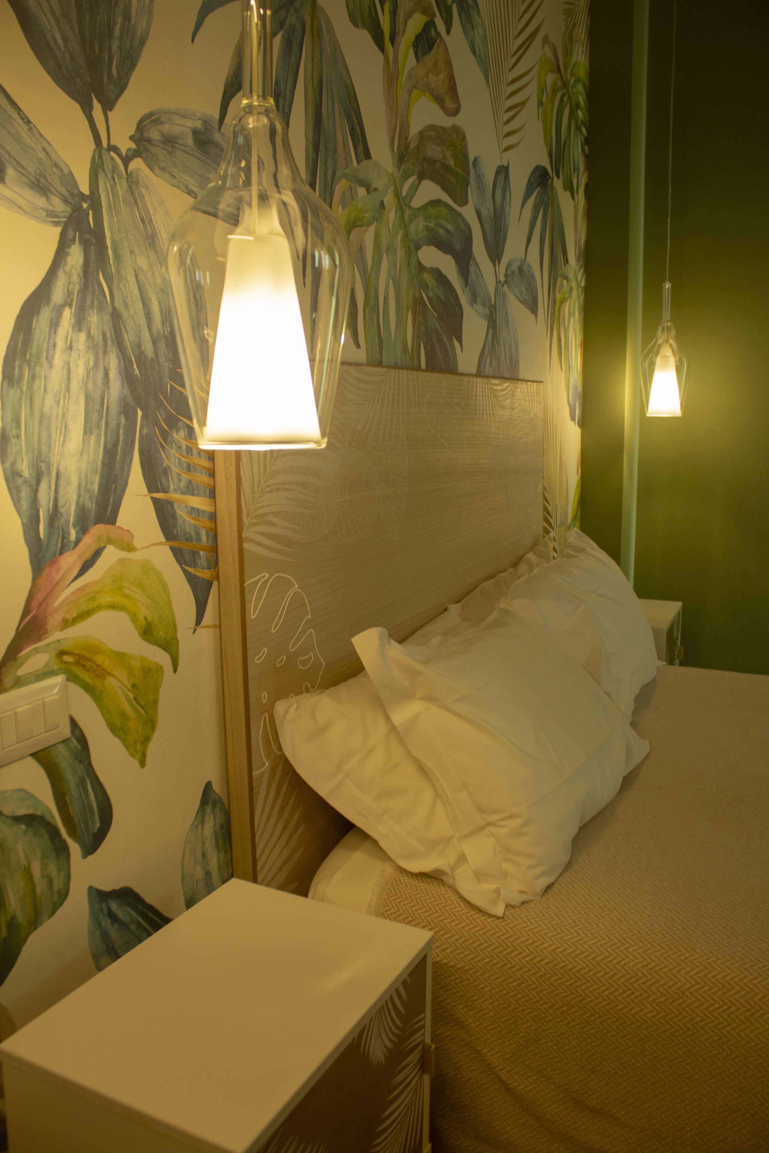 Quarto de hotel com uma cama, uma mesa de cabeceira, um abajur e uma parede com um mural de plantas tropicais.