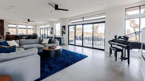 A spacious living room with a modern design, featuring a sectional sofa, a blue rug, a piano, and large windows offering a view of the outdoors.