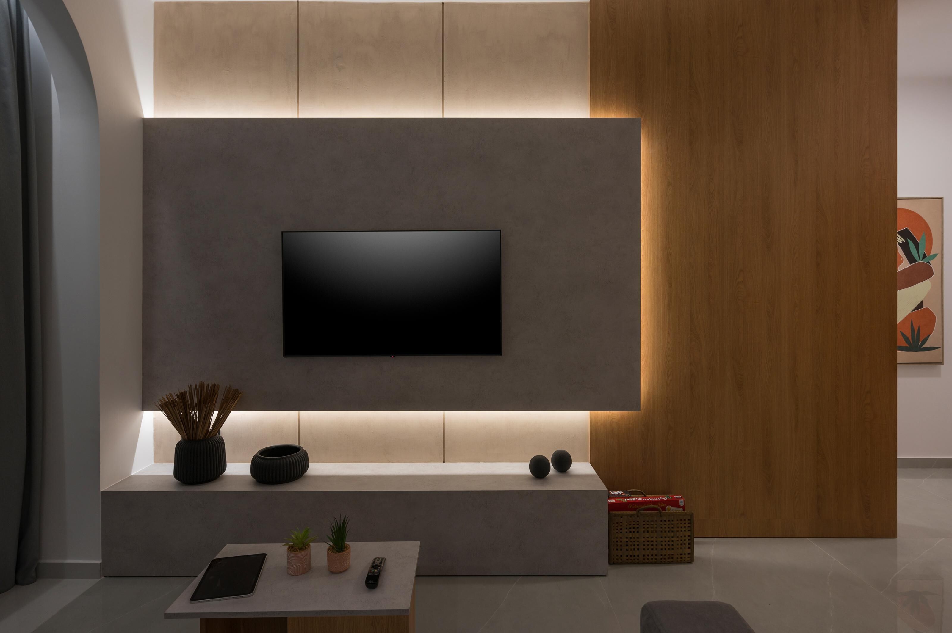 A modern living room with a flat-screen TV mounted on a wall, a low coffee table with a remote and a tablet, and a small plant on the table.