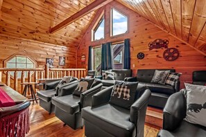 A log cabin interior with a loft bedroom, a dining area, and a seating area with leather chairs and a sofa.