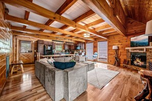 Interior acogedor de una cabaña con un gran techo de madera, chimenea, zona de cocina y espacio de estar con sofá y mesa de centro.