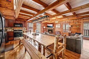 A rustic cabin interior with a wooden dining table, kitchen area, and a cozy seating area with a fireplace.