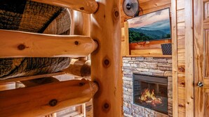 Interior acogedor de una cabaña con chimenea de piedra, paredes de madera y un televisor montado encima.