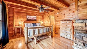 Interior de una cabaña de madera con una cama, un ventilador de techo, una chimenea de piedra y una ventana con persianas.