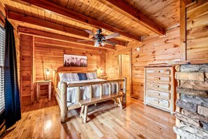 Een houten blokhut interieur met een bed, een plafondventilator, een stenen open haard en een raam met zonwering.