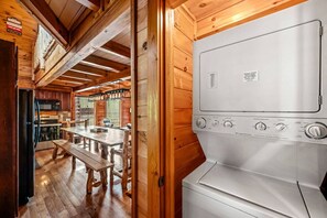 Interieur van een houten blokhut met een keukenhoek, met een grote witte kookplaat, een houten eettafel en bankjes.