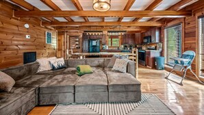 Interior acogedor de una cabaña con un amplio sofá modular, vigas de madera, una zona de cocina y una mesa de comedor.