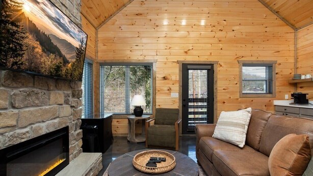 A cozy cabin interior with a stone fireplace, wooden walls, a leather sofa, and a dining area.