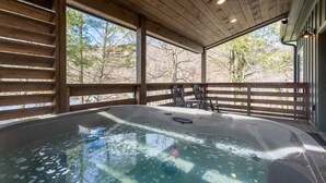 A hot tub in a wooden cabin with a view of trees and a deck with chairs.