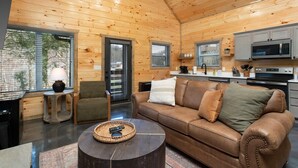 A cozy cabin interior with a wooden ceiling, a brown leather sofa, a small round coffee table, a kitchen area with modern appliances, and a window with blinds.