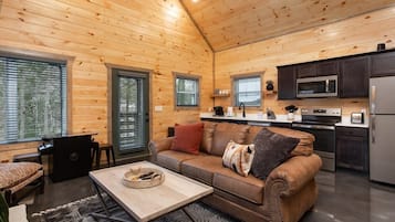 Un interior de cabaña acogedor, con techo de madera, un sofá, una mesa de centro, una zona de cocina con microondas y refrigerador, y ventanas amplias.