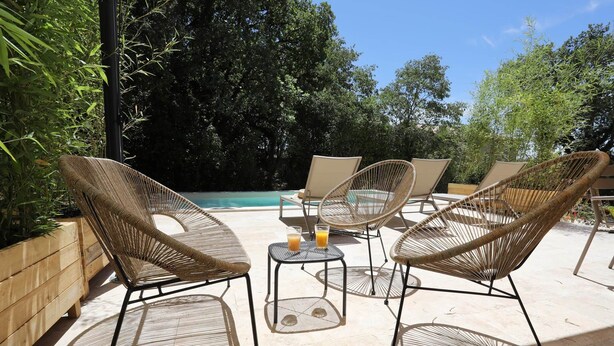 Une terrasse dotée de chaises en osier et d’une petite table où sont disposées des boissons, le tout entouré d’une végétation luxuriante et d’une piscine.