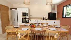 On aperçoit un coin repas avec une table en bois dressée pour six personnes, entourée de chaises en osier. Au fond, on distingue une cuisine avec des meubles blancs et un réfrigérateur noir.