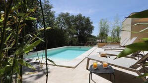 Une piscine entourée de verdure, avec des chaises longues et une table où sont servis des boissons.