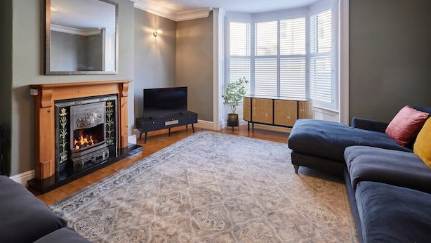 A living room with a fireplace, a television, a large window with blinds, and a dark-colored sofa.
