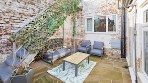 A small, enclosed patio area with a brick wall, a wooden table, grey sofas, and a potted plant.