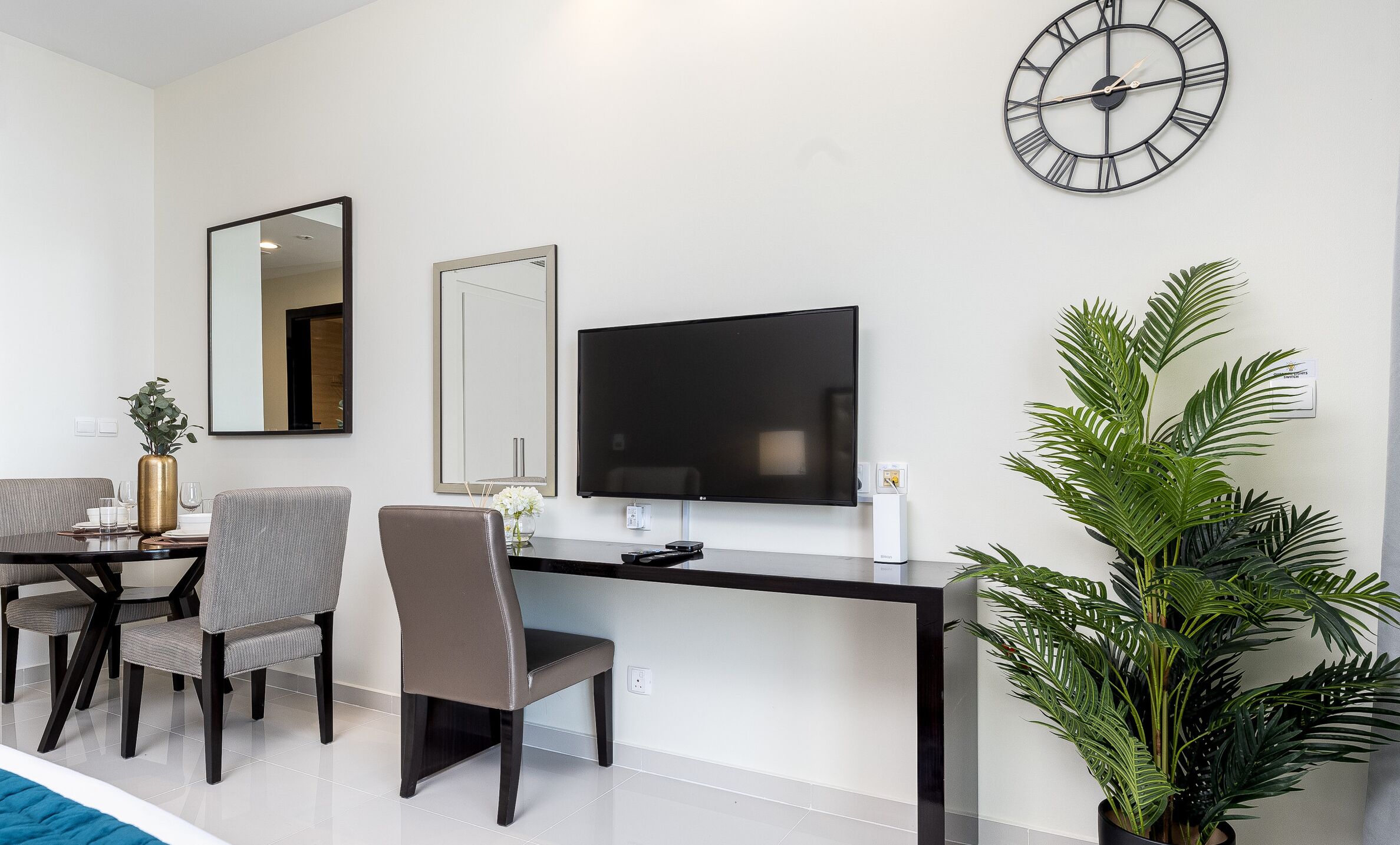 Een moderne woonkamer met een flatscreen televisie, een grote wandklok, een eethoek met een tafel en stoelen, en een potplant.