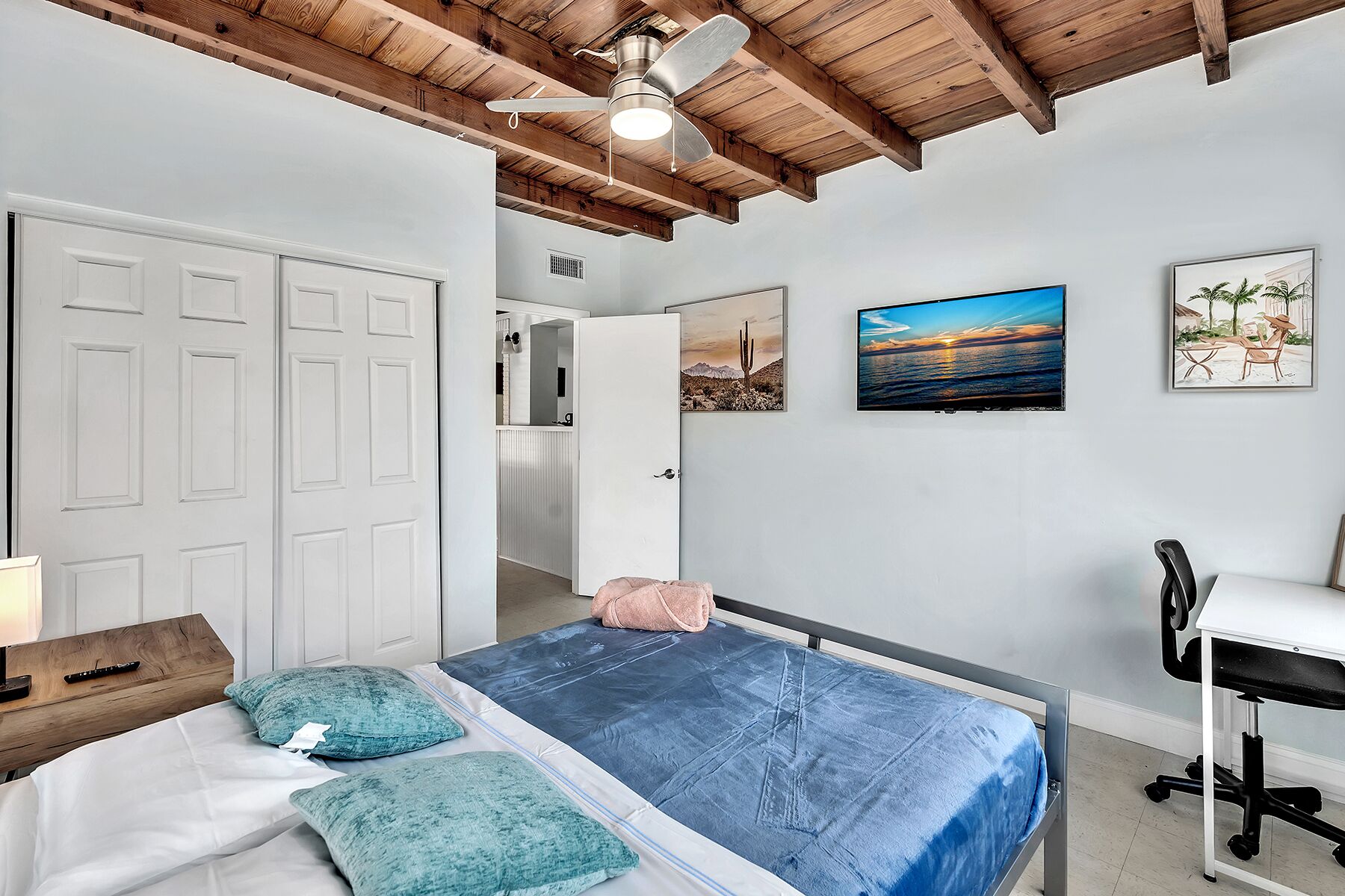 A bedroom with a bed, a desk, a chair, a ceiling fan, and artwork on the walls.