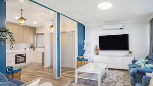 A modern living room with a white coffee table, blue sofas, a flat-screen TV, and a kitchen in the background. - The Sweet Retreat - Hot tub with Lakeviews! (Tuggerawong)