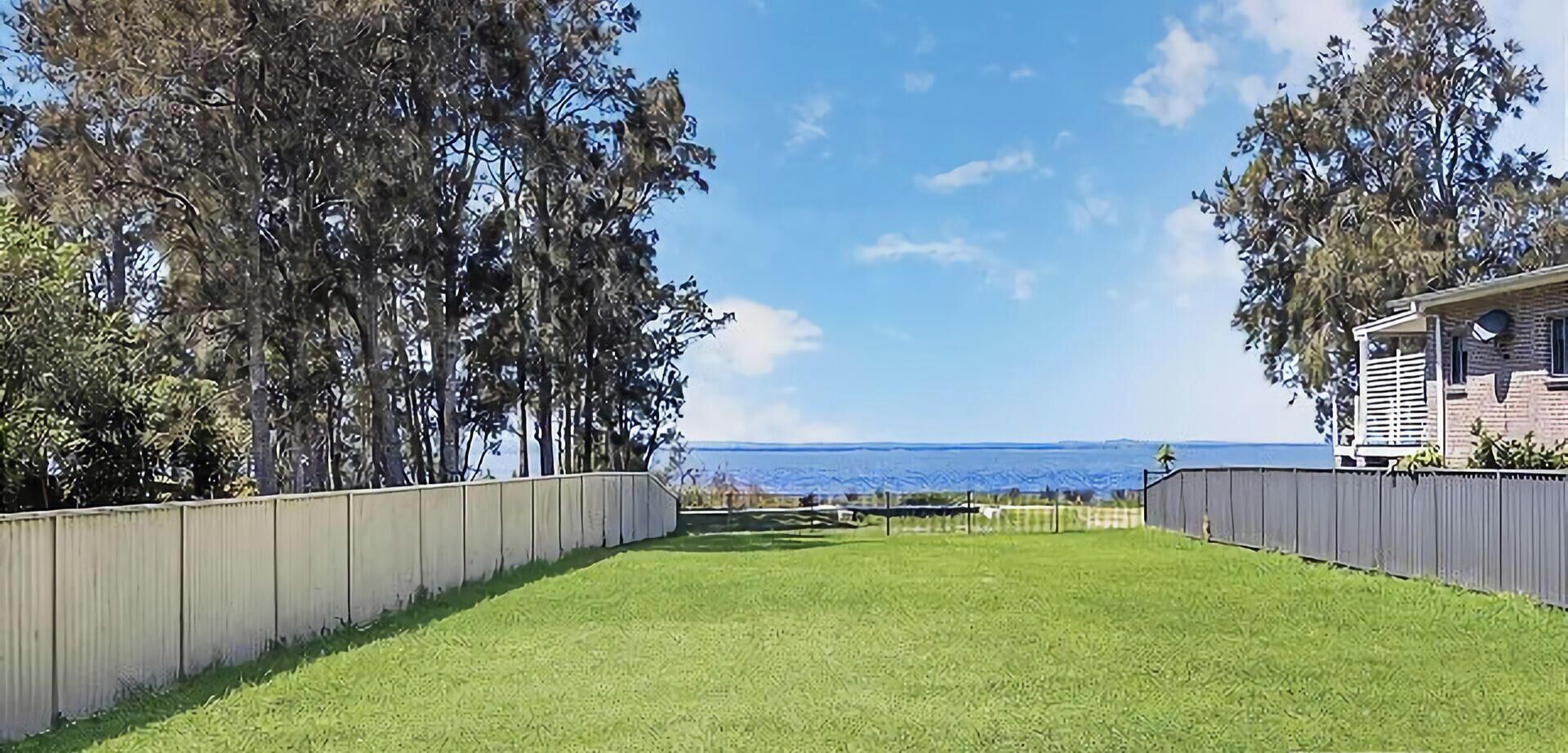 A fenced backyard with a grassy lawn and trees, leading to a house with a visible deck.