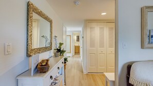 A hallway leading to a bedroom with a large mirror, a small table with a bowl of fruit, and a door with louvered panels. - Sunset Rentals | Bluff Villas 1768 (Hilton Head Island)