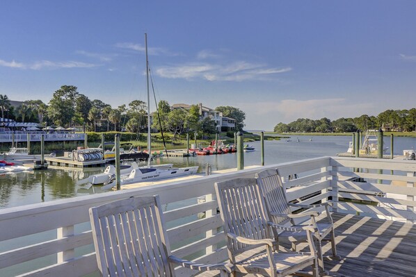 A marina with boats docked, a wooden deck with chairs, and a view of a house across the water.