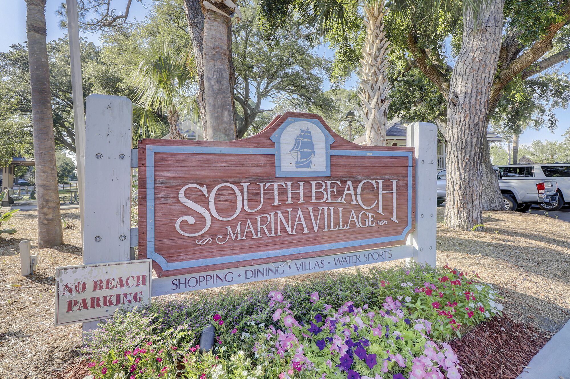 Letrero de South Beach MarinaVillage con un logotipo de barco, rodeado de árboles y una cama de flores.