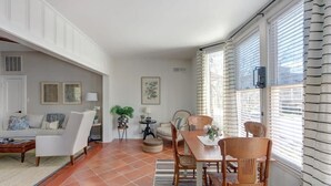 En stue med sofa, sofabord og stole, et spiseområde med bord og stole, samt et stort vindue med persienner.