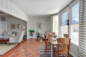 A living room with a sofa, coffee table, and chairs, a dining area with a table and chairs, and a large window with blinds. - Eichberg's Suite, Historic | Steps to Forsyth Park (Savannah)