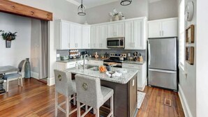A modern kitchen with white cabinets, stainless steel appliances, a granite countertop, and a central island with bar stools. - The Alfred Inn, Victorian District | Modern Flare (Savannah)