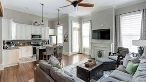 A spacious living room with a fireplace, a kitchen with white cabinets, a ceiling fan, and a television. - The Alfred Inn, Victorian District | Modern Flare (Savannah)