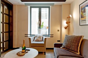 Een kamer met een houten deur, een raam met gordijnen, een plant, een fles champagne, een glas, een stoel, een bank en een lijst met een afbeelding aan de muur.