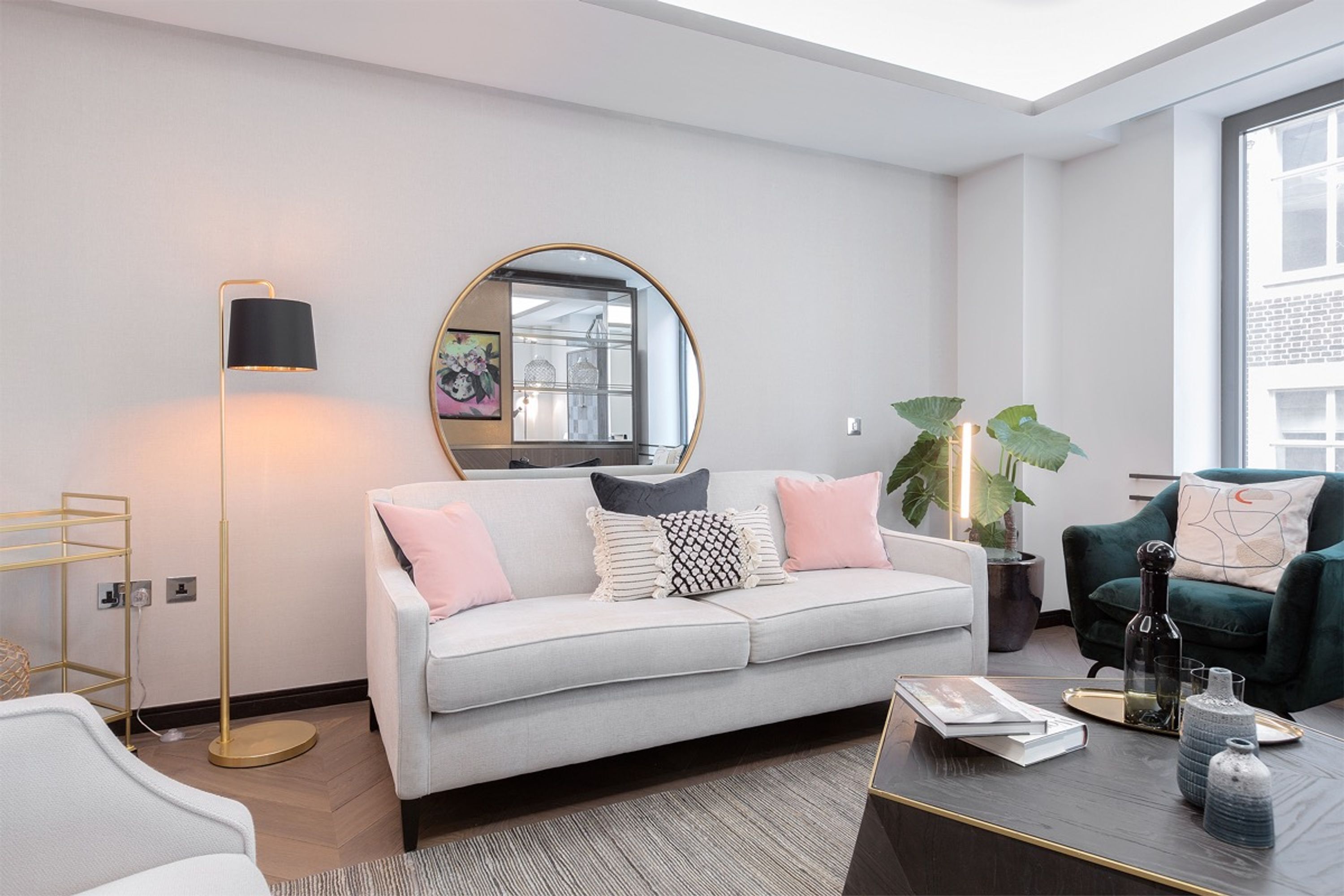A modern living room with a grey sofa, a round mirror, a floor lamp, a green armchair, and a coffee table with books and decorative items.
