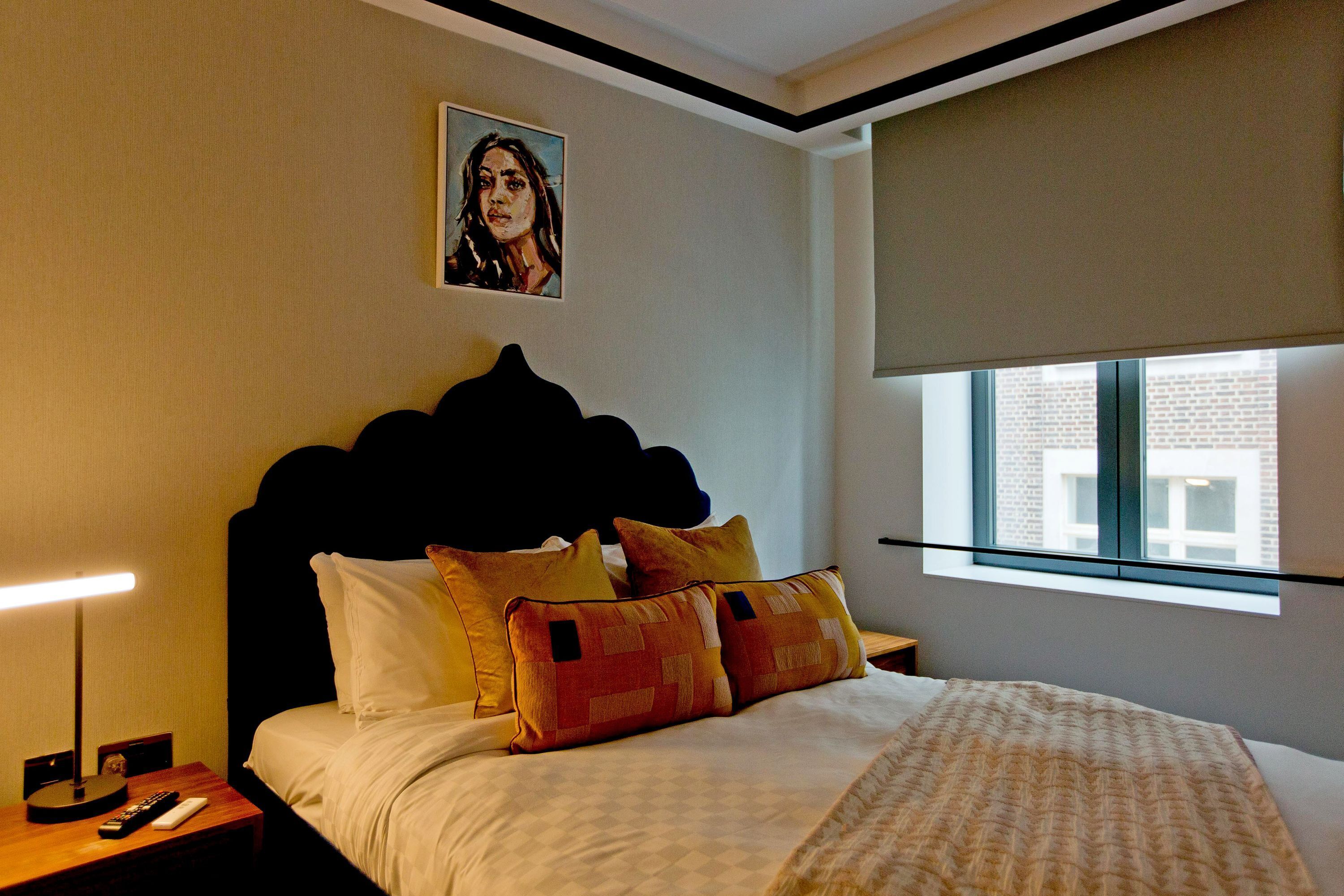 Una camera d'albergo con un letto, un comodino, una lampada, un quadro appeso al muro e una finestra vista edifici.