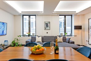 A modern living room with a wooden dining table, a bowl of fruit, a bottle of champagne, and a grey sofa with cushions. - 2-bed Superior Apt 103 (London)