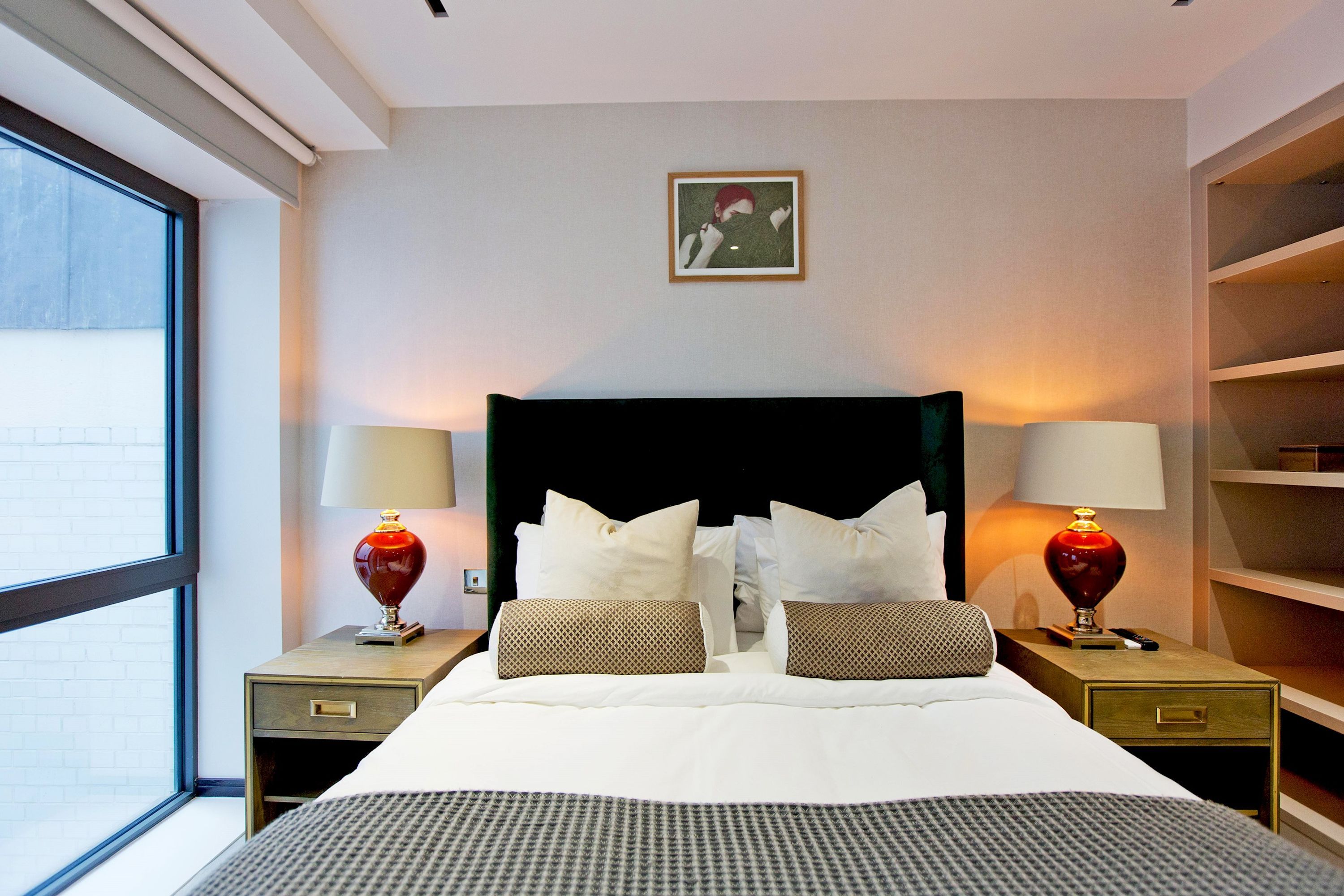 A hotel room with a large bed, two bedside tables with lamps, a framed picture on the wall, and a view through a window.