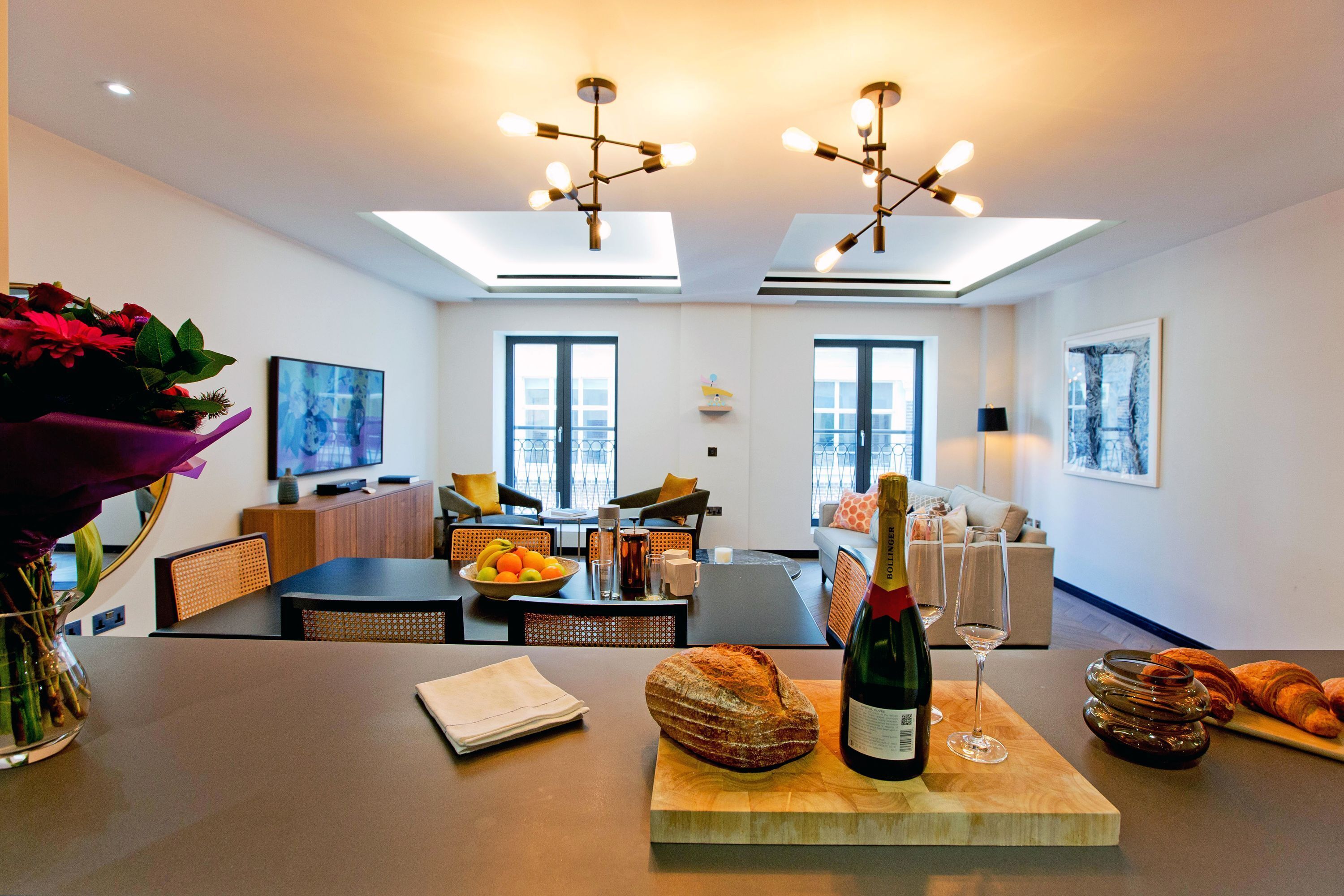 A modern living room with a dining area, featuring a wooden table with a bottle of wine, a glass, and a loaf of bread.