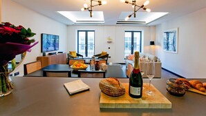 A modern living room with a dining area, featuring a wooden table with a bottle of wine, a glass, and a loaf of bread. - 2-bed Superior Apt 415 (London)